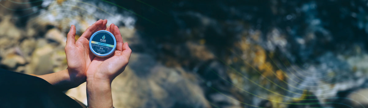 Hands holding a Pure Spectrum CBD tin beside a mountain stream